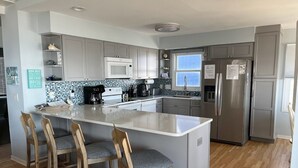 A modern kitchen with a central island, bar stools, stainless steel appliances, and a tiled backsplash. - Tropic like Its Hot Oak Island Oceanfront House, Tropic Like Its Hot Oak Island- Beach Front (Oak Island)