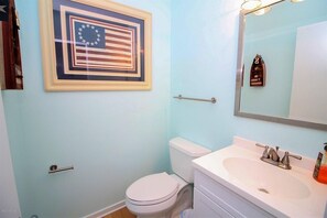 A bathroom with a light blue wall, a framed American flag, a white toilet, a sink with a mirror, and a towel rack. - Tropic like Its Hot Oak Island Oceanfront House, Tropic Like Its Hot Oak Island- Beach Front (Oak Island)