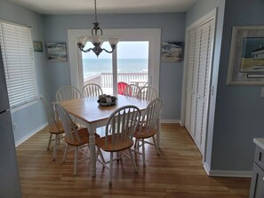 A dining area with a wooden table and white chairs, a chandelier, and a view of the ocean through a large window. - Tropic like Its Hot Oak Island Oceanfront House, Tropic Like Its Hot Oak Island- Beach Front (Oak Island)
