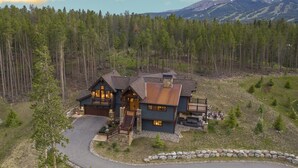 A large, two-story house with a stone foundation, a wrap-around porch, and a deck. It is surrounded by a forested area with a mountain in the background. - Spacious Elegance, 5 en suites, hiking trail to town, packed with amenities! (Breckenridge)