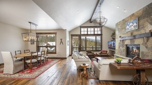 A spacious living area with a wooden floor, a fireplace, and a large window overlooking a snowy landscape. - Spacious Elegance, 5 en suites, hiking trail to town, packed with amenities! (Breckenridge)