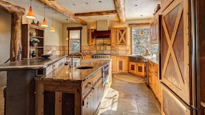 A rustic kitchen with wooden cabinets, a large island with a sink, and pendant lighting. - Rustic-elegance wrapped in custom mountain luxury, steps from Baldy Hiking Trail and shuttle stop (Breckenridge)