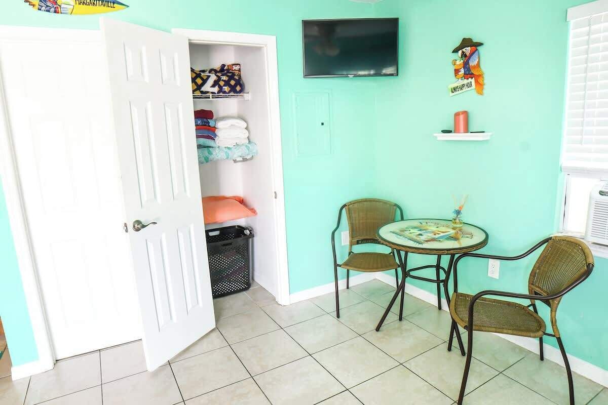 Una habitación con pared verde azulado, una puerta blanca cerrada, un televisor y una pequeña mesa con dos sillas de mimbre.