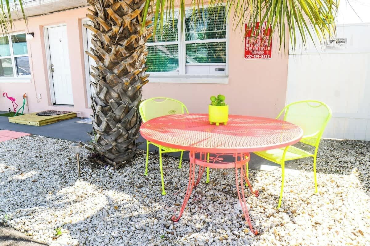 A small outdoor seating area with a round table and chairs, a palm tree, and a building in the background.