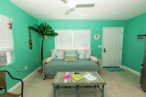 A room with a teal wall, a ceiling fan, a bed with a 'BEACH' pillow, a coffee table with papers and a phone, and a door with a doormat.