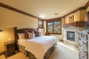 A bedroom with a large bed, a stone fireplace, and a view of a mountainous landscape through the window. - Silver Strike by Luxury Mountain Destina (Park City)