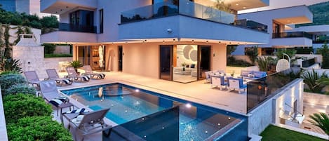 A modern two-story house with a swimming pool, outdoor seating, and a large glass door leading to the interior.