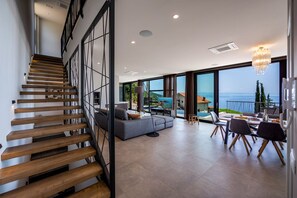 A modern interior with a staircase, a living area with a sofa and chairs, and large windows offering a view of the sea.