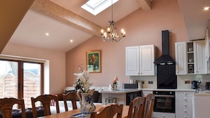 A dining area with a wooden table and chairs, a chandelier, and a kitchen with white cabinets and a black range hood. - Roosters Rest - UK31826 (Barley, near Clitheroe)