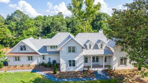 A two-story house with a symmetrical design, featuring a central entrance with a covered porch, a landscaped front yard, and a backdrop of dense trees. - 600 Linden: 8  BR, 5.5  BA House in Pinehurst, Sleeps 20 (Pinehurst)