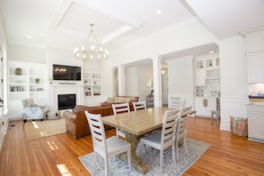 A spacious living room with a wooden dining table, white chairs, a fireplace, and a television mounted on the wall.
