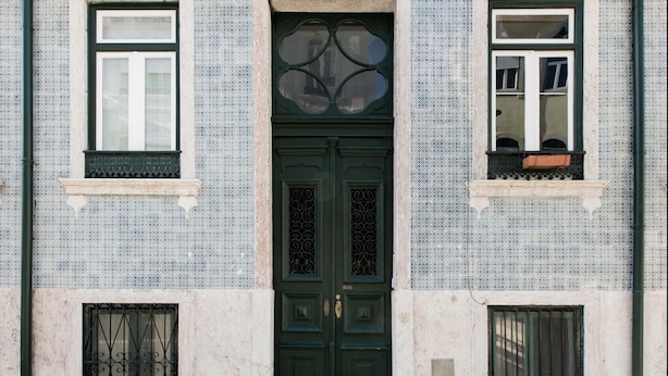 Une façade d’immeuble avec une porte verte au centre, des carreaux bleus et blancs formant un motif, et deux fenêtres dotées de balcons.