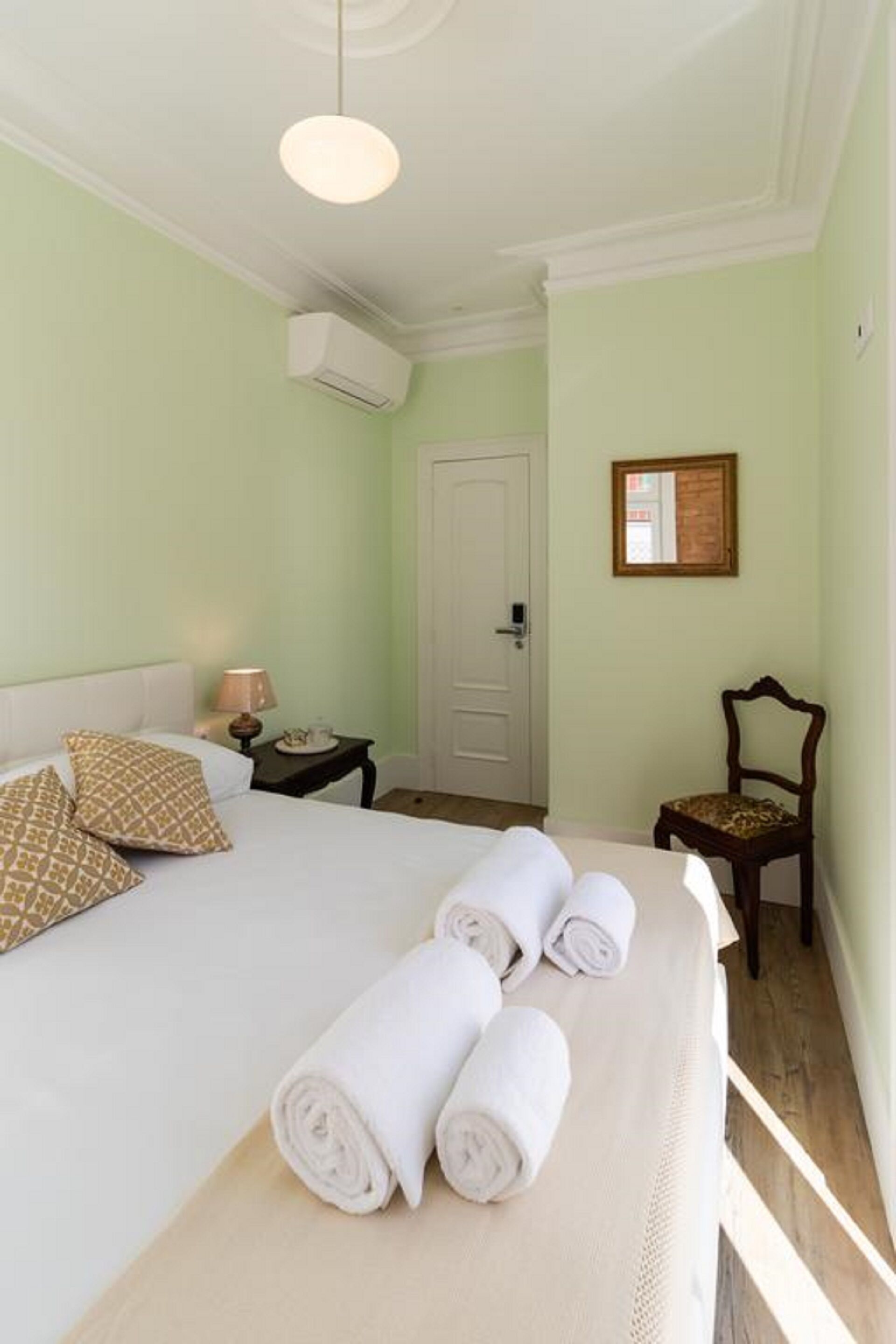 A hotel room with a bed, a nightstand, a chair, a door, and a wall-mounted air conditioning unit.