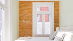 Une chambre avec un mur en briques, une fenêtre avec des volets, un lit avec une literie à rayures et une pancarte « Bienvenue ».