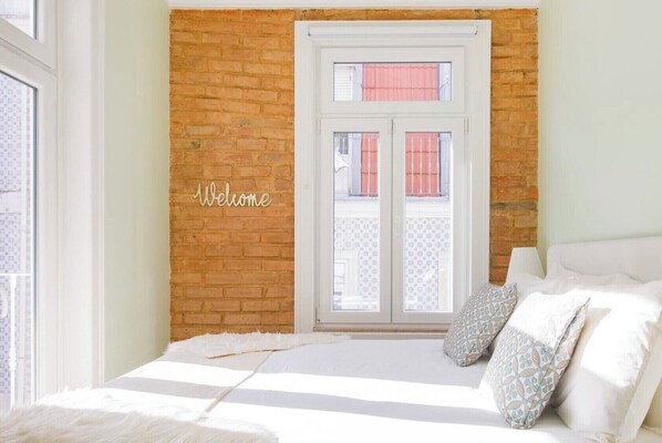 Une chambre avec un mur en briques, une fenêtre avec des volets, un lit avec une literie à rayures et une pancarte « Bienvenue ».
