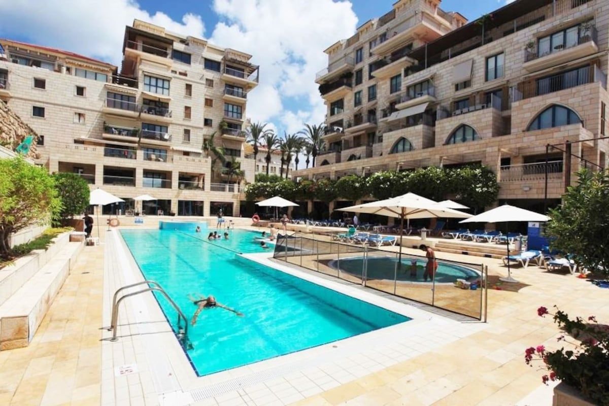 A swimming pool area with multiple pools, surrounded by buildings with balconies and outdoor seating.