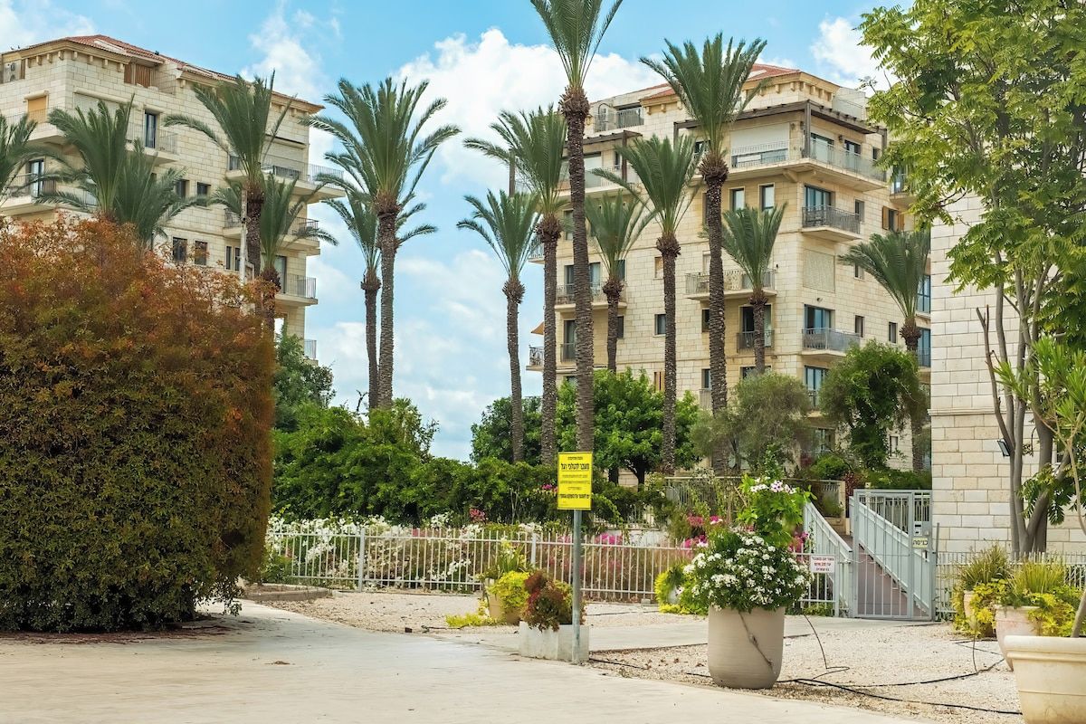 A multi-story residential building with balconies, surrounded by palm trees and a gated entrance.
