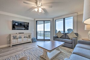 Ein Wohnzimmer mit Deckenventilator, einem Flachbildfernseher, einem grauen Sofa mit Kissen, einem Holztisch und einem großen Fenster mit Blick auf einen Balkon.