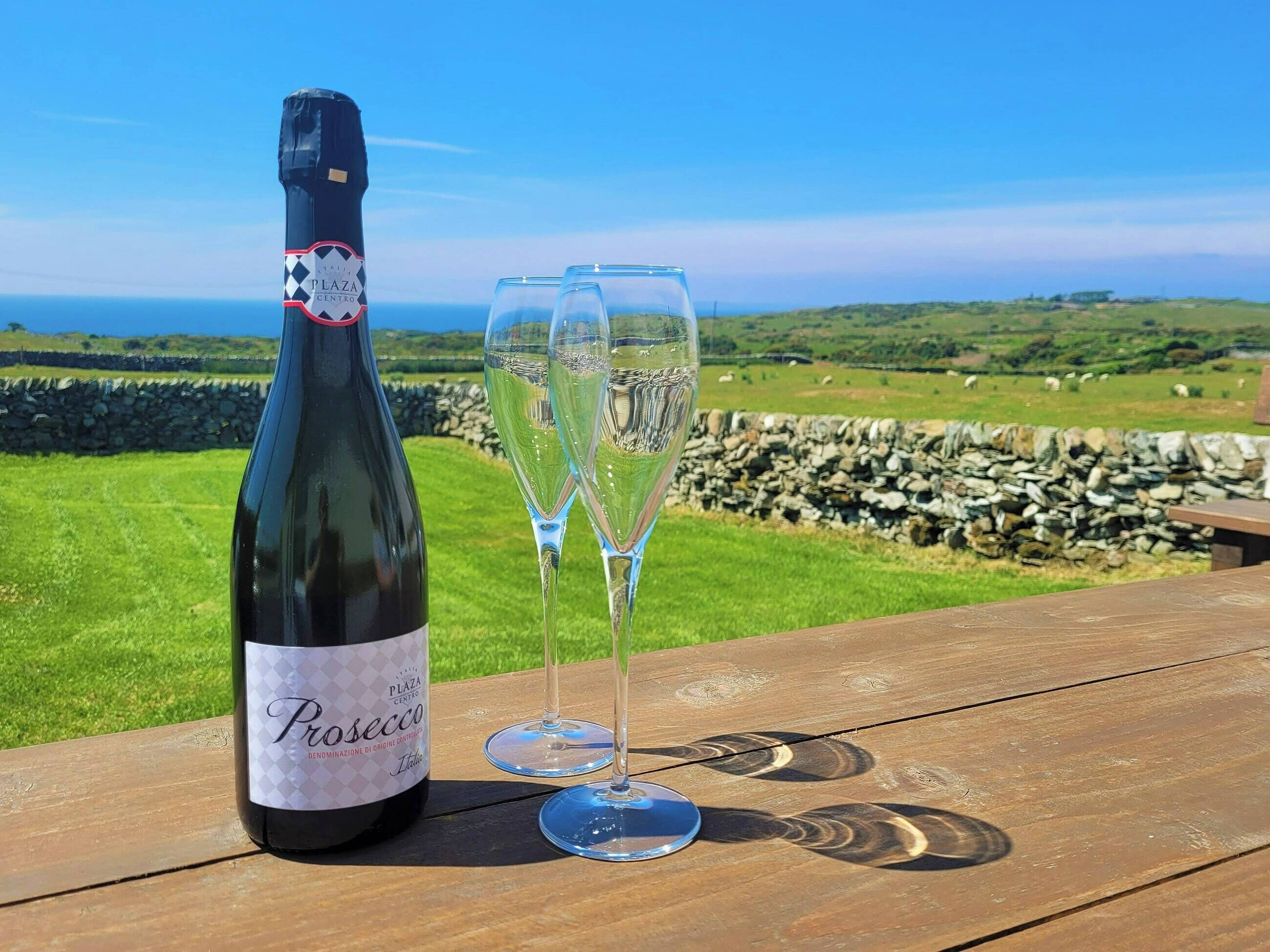 A bottle of prosecco and two glasses on a wooden table with a scenic view of green fields and a stone wall.