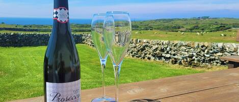 A bottle of prosecco and two glasses on a wooden table with a scenic view of green fields and a stone wall.