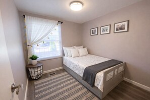 A bedroom with a bed, a window with curtains, a bedside table with a plant, and three framed pictures on the wall.