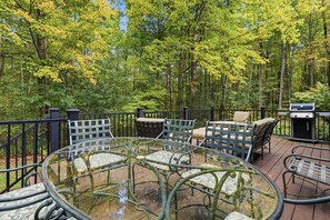 A glass-top table with chairs, a grill, and a deck surrounded by trees.