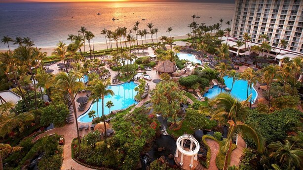 Ein Resort mit mehreren Swimmingpools, Palmen und Blick auf den Ozean im Sonnenuntergang.