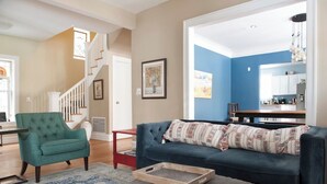 A living room with a green armchair, a blue sofa, and a grey ottoman. There is a staircase in the background leading to another room. - The Greenleaf House—Whole House (Chicago)