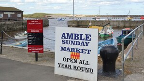 Placa indicando o Mercado Domingo de Amble, aberto o ano todo, com barcos atracados por perto.