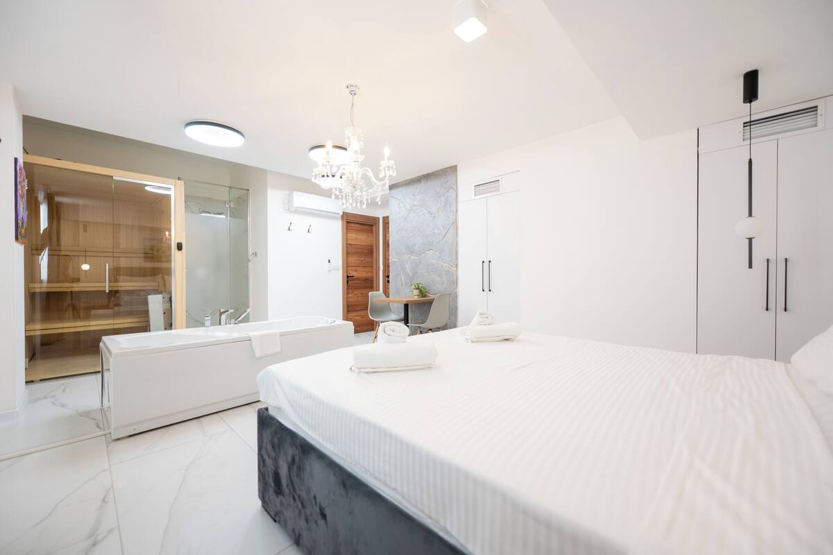 A modern bedroom with a large bed, a walk-in closet, a bathroom with a glass shower, and a chandelier.