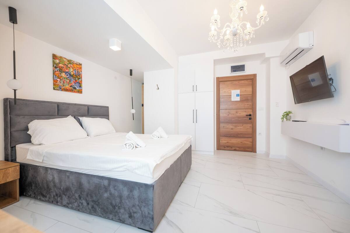 A modern bedroom with a large bed, a chandelier, a TV, and a wooden door.