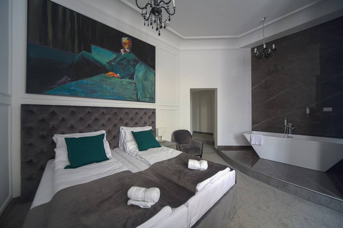 A modern bedroom with a large bed, a painting, a chandelier, a bathroom with a bathtub, and a marble wall.