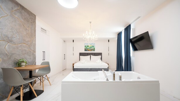 A modern bedroom with a large white bathtub, a bed with white linens, a dining table with chairs, and a TV mounted on the wall. - Kings Palace Suites (Belgrade)
