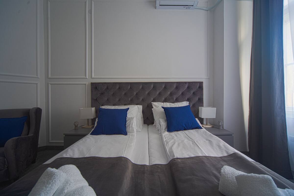 A hotel room with a double bed, two nightstands with lamps, a tufted headboard, and a large window with curtains.