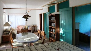 A cozy living room with a round glass table, wooden chairs, a bookshelf, and a teal door. - Casa Pájaros - Entire Lower Unit (Cabo Pulmo)