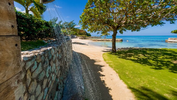 A stone wall with a water feature, a pathway leading to a beach, and a tree with a view of the ocean. - A beautiful private beachfront paradise in Old Fort Bay, Ocho Rios (St. Ann Parish)