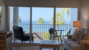 Ein Wohnzimmer mit einem großen Schiebeglasfenster, das auf einen Balkon mit Blick auf das Meer und Palmen führt.