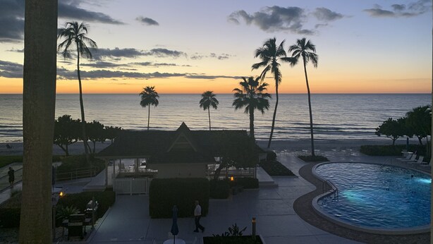 Ein Poolbereich eines Resorts mit Blick auf den Ozean bei Sonnenuntergang, Palmen und eine Lounge mit Sitzgelegenheiten im Freien.
