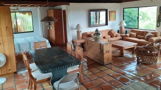 Une pièce chaleureuse avec un toit de chaume, une table à manger en bois entourée de chaises, un ensemble de canapés et une fenêtre donnant sur un paysage verdoyant.