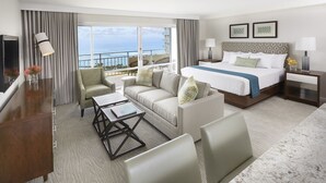 Een hotelkamer met een bed, een bank, een bureau en een balkon met uitzicht op de oceaan.