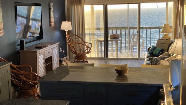 A room with a kitchen area, a dining table with chairs, a large window with a view of the ocean, and a ceiling fan. - Renovated 2/2 Gulf Front Condo (Indian Shores)