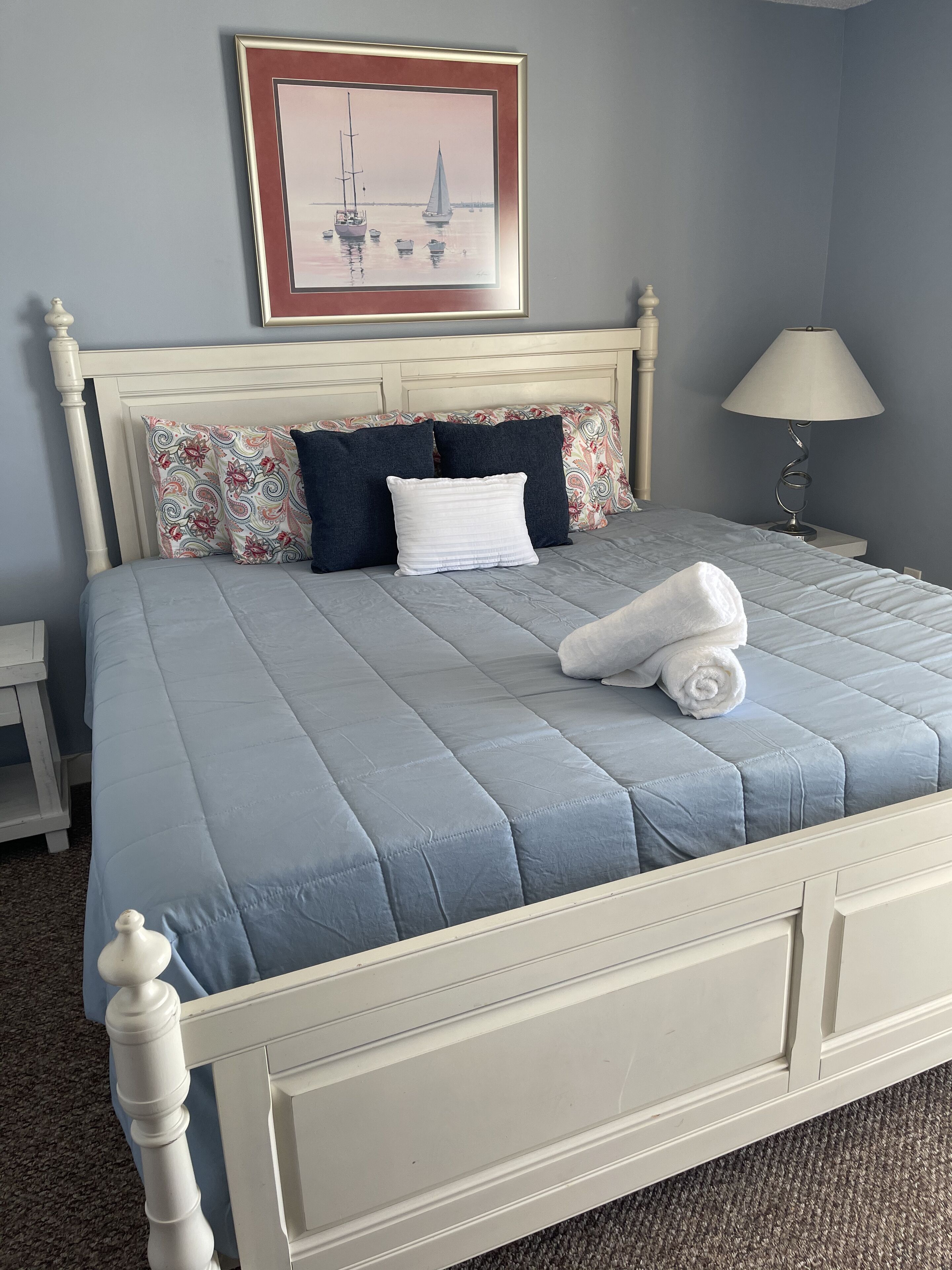 A neatly made bed with a blue bedspread, white headboard, and decorative pillows. A framed artwork hangs above the bed, and a bedside lamp is visible.