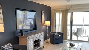 A modern living room with a flat-screen TV, a ceiling fan, a dining area with a glass-top table, and a balcony with outdoor furniture. - Renovated 2/2 Gulf Front Condo (Indian Shores)