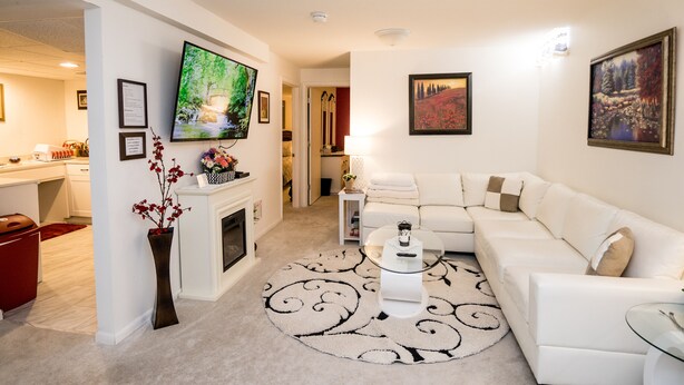 A modern living room with a white sofa, a fireplace, a flat-screen TV, and artwork on the walls. - Hutchinson Haven of Rest (Hutchinson)