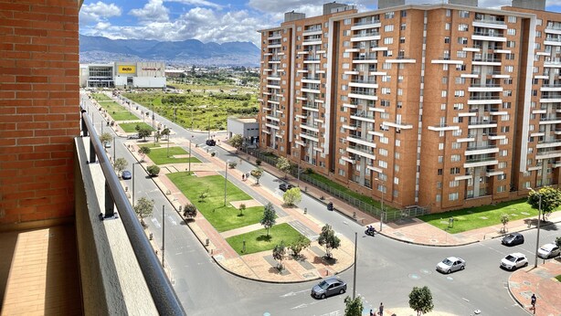 A high-rise residential building with multiple balconies, a well-maintained green space, and a clear view of a distant cityscape. - WOW! NEW 3/2BR, NEAR AIRPORT & SHOPPING - PREMIUM! (Bogotá)