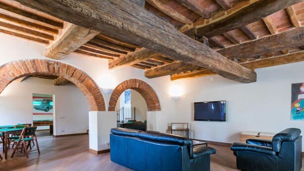 Uma sala ampla com vigas de madeira aparente, um sofá azul, um estofado de couro preto e uma área de jantar com mesas e cadeiras de madeira.