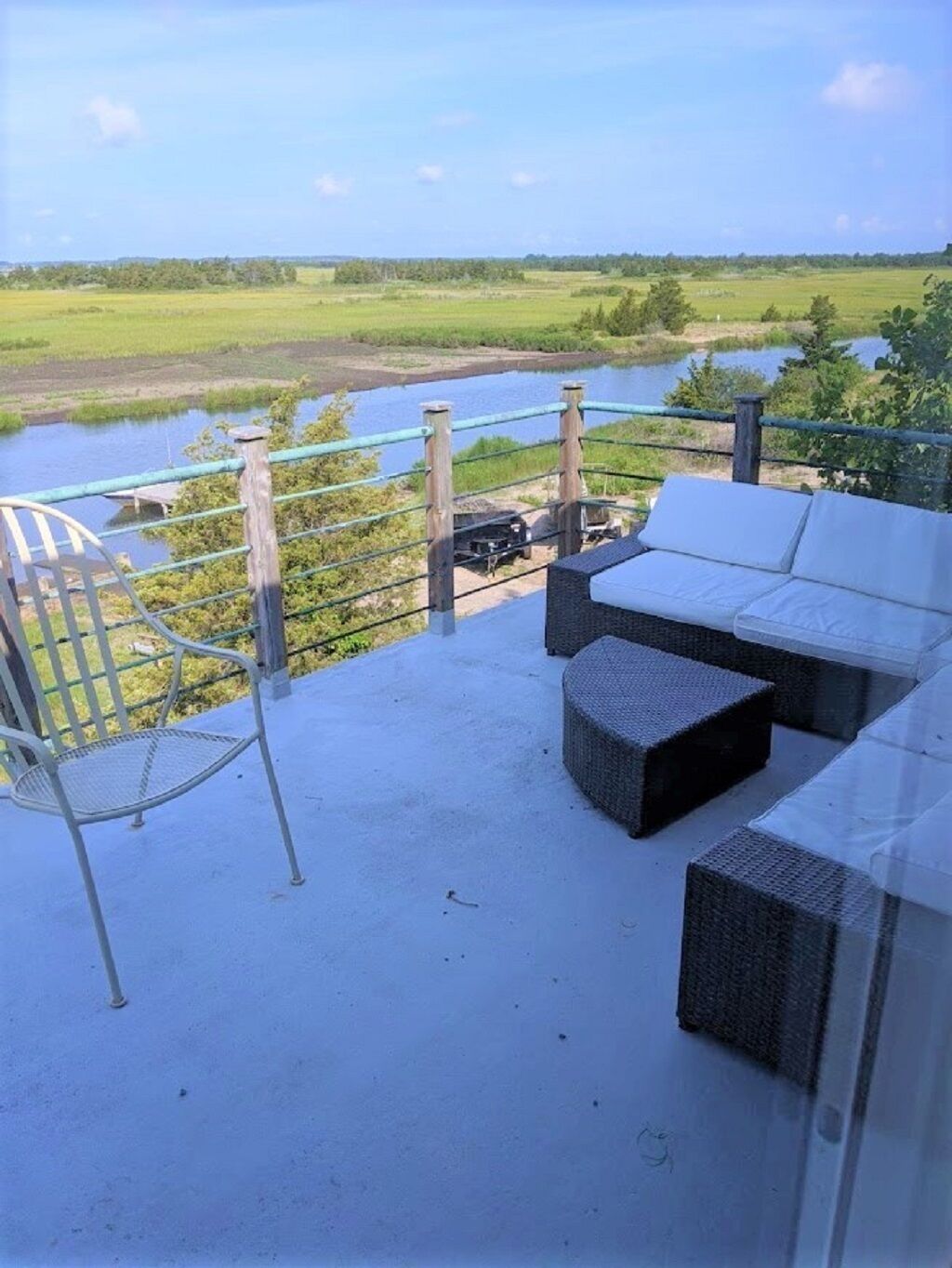 Un balcon avec vue sur une rivière et des champs, aménagé avec un canapé blanc et des fauteuils en osier.