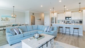A modern living room with a light blue sectional sofa, a coffee table, and a kitchen area with white cabinets and bar stools.