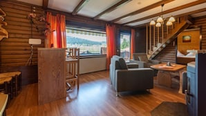 Accogliente interno di una baita in legno, con zona pranzo in legno, un divano, un tavolino e una scala che conduce a una camera da letto soppalco.