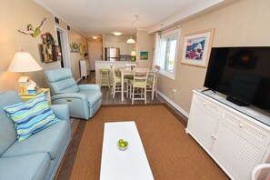A living room with a sofa, a coffee table, a TV, and a dining area with chairs and a table. - Beach House 309 - Oceanfront in Garden City Beach, SC (Garden City)